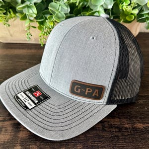 May include: A heather gray trucker hat with a black mesh back and a flat brim. The front features a rectangular brown leather patch with the text G-PA engraved in black. A black and white sticker on the brim reads Richardson 112 Trucker Adjustable.