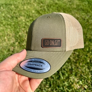 May include: Olive green and tan trucker hat with a leather patch that reads "GO ON, GIT." The hat features a snapback closure and a Yupoong tag. The hat is a classic accessory.