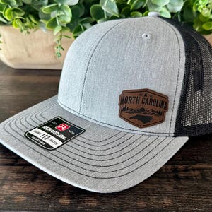 May include: A gray and black trucker hat with a curved brim. The hat features a brown leather patch with the words "North Carolina" and a state outline. The hat is adjustable and has a Richardson 112 Trucker tag.