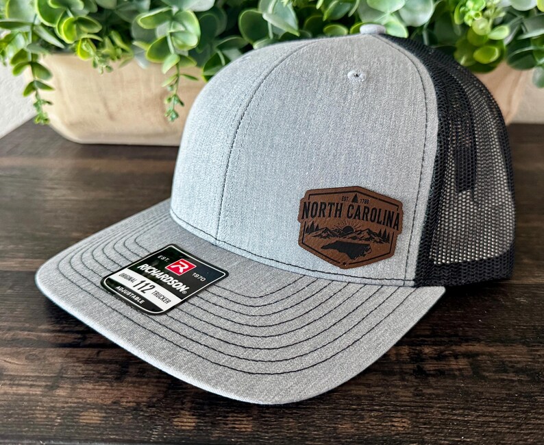 May include: A gray and black trucker hat with a curved brim. The hat features a brown leather patch with the words "North Carolina" and a state outline. The hat is adjustable and has a Richardson 112 Trucker tag.