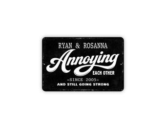 May include: A rectangular black metal sign with white text that reads RYAN & ROSANNA, Annoying EACH OTHER, -SINCE 2005-, AND STILL GOING STRONG. The sign features a distressed texture and rounded corners, with a bold, stylized script font for the word Annoying.