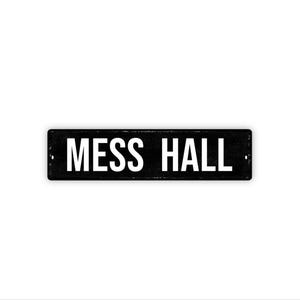 May include: A rectangular black metal sign with the words "MESS HALL" in bold white capital letters. The sign has a distressed appearance and is mounted on a white background.