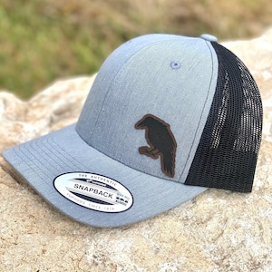 May include: A light blue Snapback baseball cap with a black mesh back and a curved brim. The cap features a brown leather patch with a raven design. The tag reads "The Authentic Snapback Yupoong Since 1974".