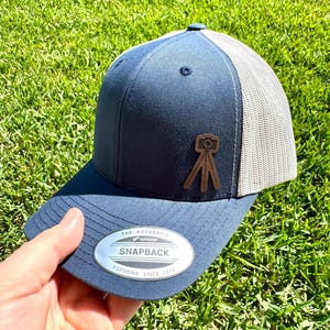 May include: Navy blue snapback hat with a gray mesh back. The hat features a brown leather patch with a camera design. The hat has the text "The Authentic Snapback" and "Yupoong Since 1974" on the front.