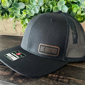 May include: Black trucker hat with a mesh back and a curved brim. The hat features a rectangular patch that reads "BIG BROTHER" in a copper color. The hat is sitting on a dark wooden surface.
