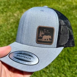 May include: A gray and black trucker hat with a brown leather patch featuring a bear, trees, and mountains. The hat has a mesh back and a curved brim. The tag reads "The Authentic Snapback Yupoong Since 1974."