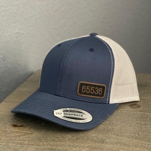 May include: A navy blue and white trucker hat with a leather patch displaying the number "65536". The hat features a curved brim and a snapback closure. The text "THE AUTHENTIC SNAPBACK YUPOONG SINCE 1974" is printed on a tag.