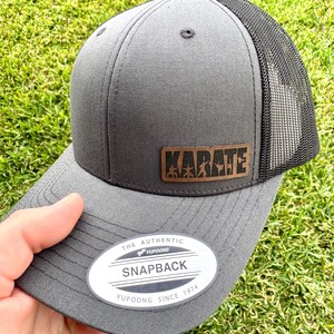 May include: Gray snapback hat with a black mesh back. The hat features a brown leather patch with the word "KARATE" and a graphic. The hat has a curved brim and a white tag that reads "SNAPBACK".