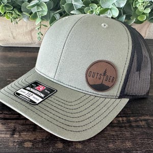 May include: A green and black trucker hat with a curved brim. The hat features a circular patch with the word "OUTSIDER" and a tree graphic. The hat has a Richardson 112 Trucker tag.