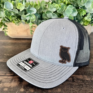 May include: Gray trucker hat with a black mesh back and a curved brim. The hat features a brown leather patch with a black owl design. The Richardson 112 adjustable hat has a structured front and a snapback closure.