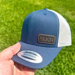 May include: Navy blue and white trucker hat with a curved brim. The hat features a brown leather patch with the text "PSALM 23:4". The hat also has a white mesh back and a snapback closure.