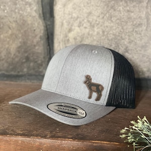 May include: A gray and black snapback hat with a brown leather patch featuring a ram silhouette. The hat has a curved brim and a mesh back. The text "THE AUTHENTIC SNAPBACK YUPOONG SINCE 1974" is visible on the front.
