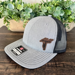 May include: Gray trucker hat with a black mesh back and a brown leather patch. The patch features a tree design. The hat has a curved brim and an adjustable strap. The Richardson brand label is visible.