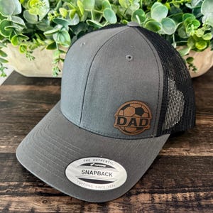 May include: A gray trucker hat with a black mesh back and a brown leather patch featuring a soccer ball graphic and the text DAD. The brim has a silver and black sticker that reads THE AUTHENTIC YUPOONG SNAPBACK SINCE 1974.