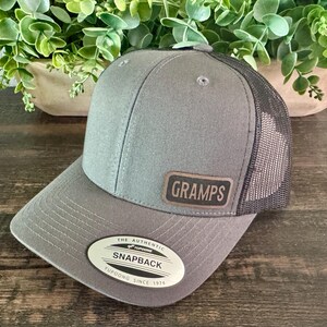 May include: Gray Snapback hat with a mesh back and a curved brim. The hat features a leather patch with the word "GRAMPS" and a white oval tag that reads "THE AUTHENTIC SNAPBACK YUPOONG SINCE 1974".