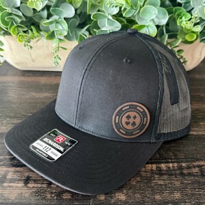 May include: Black trucker hat with a mesh back and a curved brim. The hat features a brown leather patch with a poker chip design. The Richardson 112 adjustable hat is displayed on a dark wood surface.