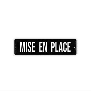 May include: A rectangular black metal sign with the words "MISE EN PLACE" in white, centered on the sign. The sign has a slightly distressed appearance, with visible wear and tear. The background is plain white.