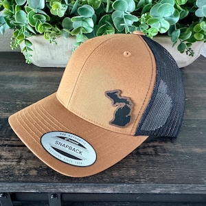 May include: A brown snapback hat with a black mesh back. The hat features a patch of the state of Michigan. The hat has a curved brim and a snap closure. The tag reads "The Authentic Yupoong Since 1974".