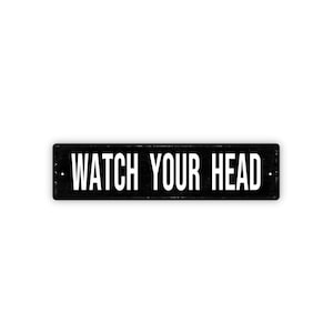 May include: A rectangular black sign with the words "WATCH YOUR HEAD" in white, centered on the sign. The sign has a distressed look, with small white dots on either side of the text.