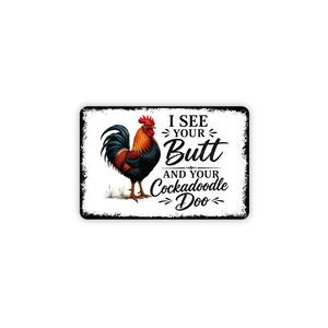 May include: A rectangular metal sign with a white background and a distressed black border. It features a colorful rooster illustration on the left and the black text I SEE YOUR Butt AND YOUR Cockadoodle Doo on the right side of the sign.
