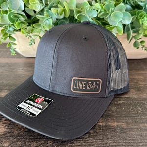 May include: Black trucker hat with a mesh back and a curved brim. The hat features a rectangular patch with the text "LUKE 15:4-7". The hat is adjustable and has a Richardson 112 tag.