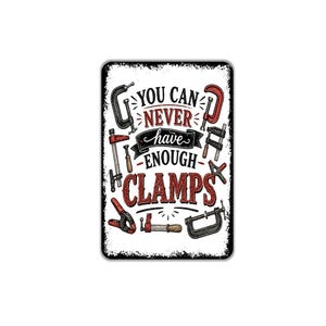 May include: A rectangular metal sign with a white background and distressed black edges. It features various illustrations of woodworking clamps in red, black, and grey, surrounding the text: YOU CAN NEVER have ENOUGH CLAMPS in black and red.