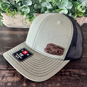 May include: An olive green baseball cap with a black mesh back and a brown leather patch featuring a fire truck graphic. The cap has a black brim with stitching and a black sticker on the brim that reads 112 The Original MD-LG.