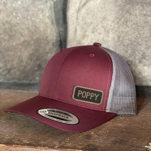 May include: A burgundy and gray snapback hat with the word "POPPY" on a rectangular patch. The hat features a curved brim and a mesh back. The tag reads "THE AUTHENTIC SNAPBACK YUPOONG SINCE 1974".