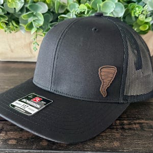 May include: Black Richardson 112 trucker hat with a brown leatherette tornado patch. The hat has a mesh back and an adjustable snap closure. The Richardson label is visible on the front.