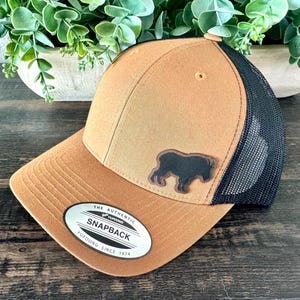 May include: A brown and black snapback hat with a black buffalo patch. The hat has a mesh back and a curved brim. The hat has a tag that reads "The Authentic Snapback Yupoong Since 1974."