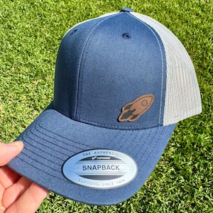 May include: A navy blue Snapback hat with a gray mesh back and a brown rocket ship patch. The hat has a curved brim and a snap closure. The text "THE AUTHENTIC YUPOONG SNAPBACK YUPOONG SINCE 1974" is on the front.