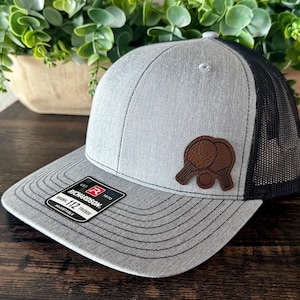 May include: Gray trucker hat with a black mesh back and a curved brim. The hat features a brown leather patch with a ping pong paddle and ball design. The tag reads "Richardson 112 Trucker".