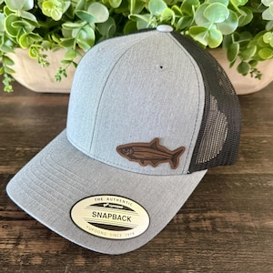 May include: Gray and black trucker hat with a fish patch. The hat features a gray front panel, a black mesh back, and a brown leather patch with a fish design. The hat has a curved brim and a snapback closure. The tag reads "THE AUTHENTIC SNAPBACK YUPOONG SINCE 1974."