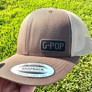 May include: A brown and tan trucker hat with the text "G-POP" on a black patch. The hat has a curved brim and a mesh back. The hat has a snapback closure and a Yupoong tag.