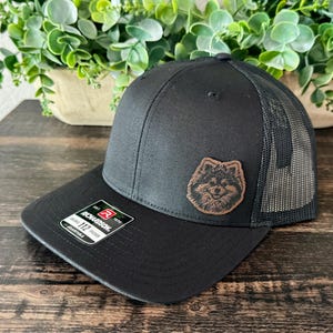 May include: A black trucker hat featuring a mesh back and a brown leather-like patch on the front panel. The patch displays an engraved illustration of a dog's face. A rectangular sticker on the brim reads Richardson 112 Adjustable.