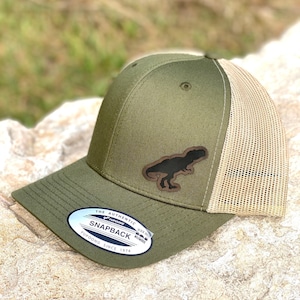 May include: Olive green and tan trucker hat with a black Tyrannosaurus Rex patch. The hat features a curved brim, a mesh back, and a snapback closure. The hat has a small, oval sticker on the brim that reads "The Authentic Snapback".