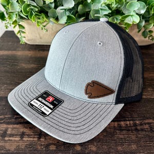 May include: A gray and black trucker hat featuring a heathered gray front panel with a brown leather arrowhead patch. The hat has a black mesh back, a flat brim with black stitching, and a black sticker on the brim that reads Richardson 112.
