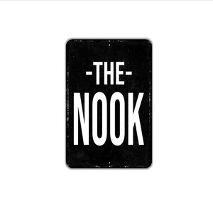 May include: A rectangular black metal sign with rounded corners featuring the white text -THE- NOOK centered on the surface. The sign has a slightly distressed texture and small mounting holes at the top and bottom center edges of the frame.