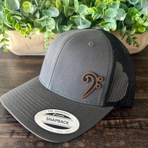 May include: A gray snapback hat with a black mesh back and a brown treble clef emblem. The hat has a curved brim and a white tag that reads "The Authentic Snapback Yupoong Since 1974".