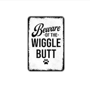 May include: A rectangular metal sign with a distressed white background and black text that reads Beware -of the- WIGGLE BUTT, featuring a small black paw print icon centered at the bottom. The edges of the sign have a worn, weathered appearance.