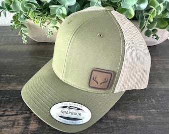 Puede incluir: Gorra snapback verde oliva con parte trasera de malla beige y un parche de cuero marrón con un diseño de cuernos de ciervo. La gorra tiene una visera curva y una etiqueta blanca que dice "The Authentic Snapback Yupoong Since 1974."