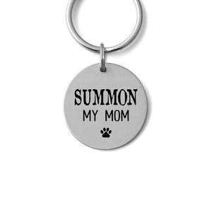 May include: A silver-colored keychain with a round tag. The tag has the words "SUMMON MY MOM" in black, with a paw print below. The keychain ring is also silver.