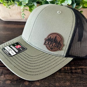 May include: Olive green and black trucker hat with a patch that says "Explore More". The hat has a curved brim and a mesh back. The hat is adjustable and has a Richardson 112 tag.