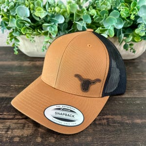 May include: A brown and black snapback hat with a leather patch of a longhorn steer. The hat has a mesh back and a curved brim. The tag on the front reads "The Authentic Snapback Yupoong Since 1974."