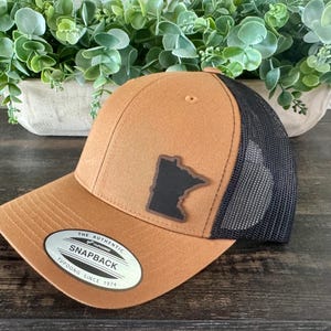 May include: A brown and black snapback hat with a black mesh back. The hat features a black leather patch with the outline of the state of Minnesota. The hat has a curved brim and a tag that reads "The Authentic Snapback Yupoong Since 1974."