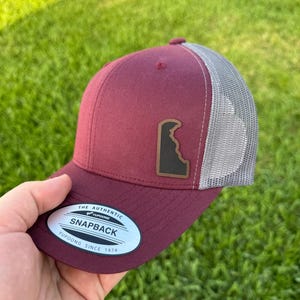 May include: A burgundy and gray snapback hat with a brown patch featuring a state outline. The hat has a curved brim and a mesh back. The tag reads "The Authentic Snapback Yupoong Since 1974."