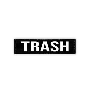 May include: A rectangular black sign with the word "TRASH" in bold white capital letters. The sign has a slightly distressed appearance, suggesting a vintage or industrial aesthetic. The background is plain white.
