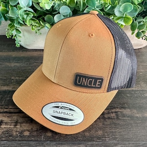 May include: A brown snapback hat with a black mesh back. The hat features a leather patch with the word "UNCLE" and a white and black oval patch that reads "SNAPBACK". The hat is a Yupoong brand.