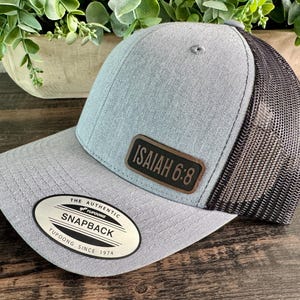 May include: A gray snapback trucker hat with a black mesh back. The hat features a leather patch with the text "ISAIAH 6:8". The brim has a curved design, and the hat has a "Yupoong Snapback" label.