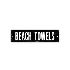 May include: A rectangular black sign with the words "BEACH TOWELS" in bold white capital letters. The sign has a distressed look, with visible texture and small holes at each end. The sign is centered on a white background.